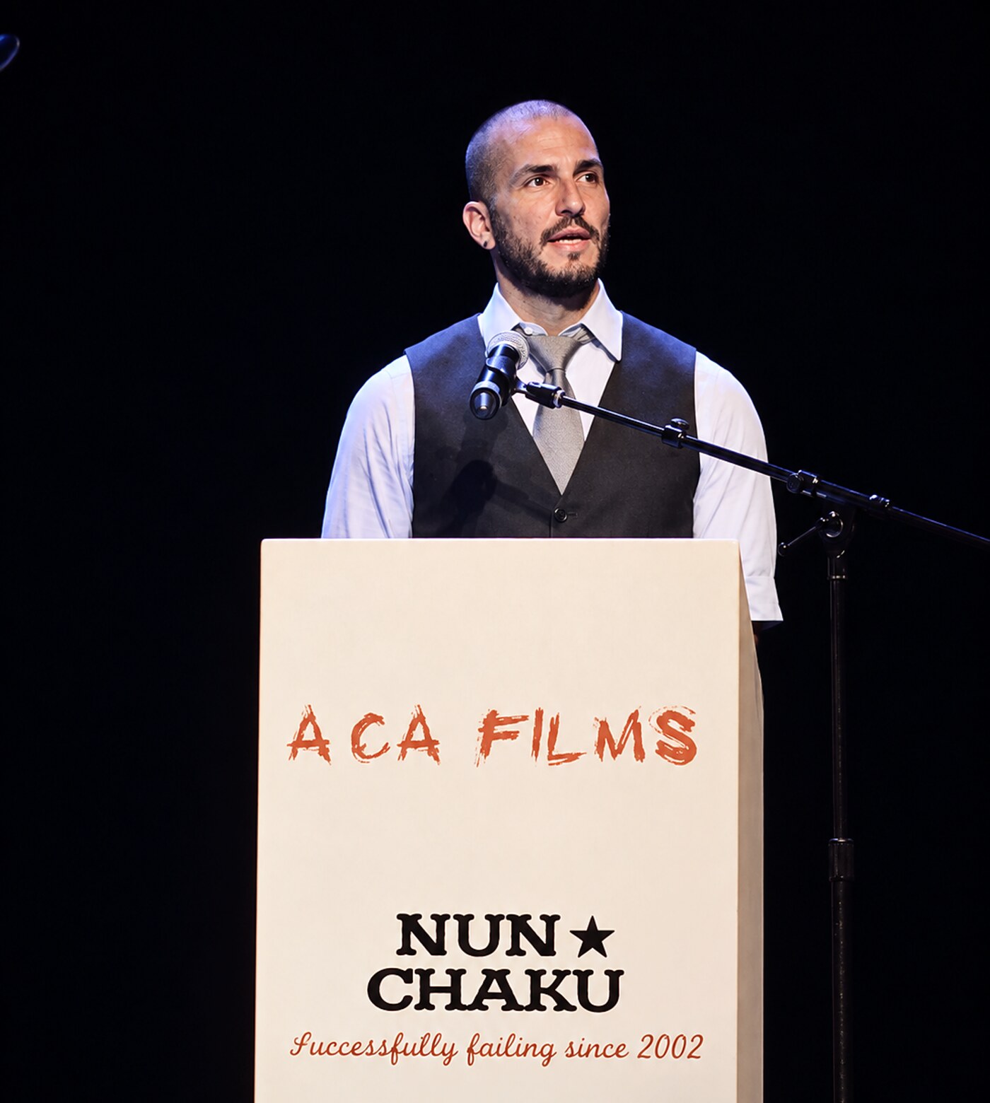 Troy Jason Valls speaking at a lectern on a dark stage in a vest and tie